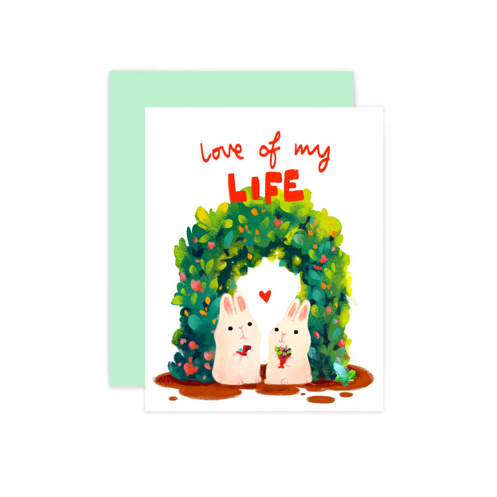 Text: Love of my Life. Illustration features a pair of white bunnies beneath a leafy green archway. One bunny holds a bouquet of flowers, the other holds a ring in a box. There is a small red heart floating above them. Text in red lettering above. White background.